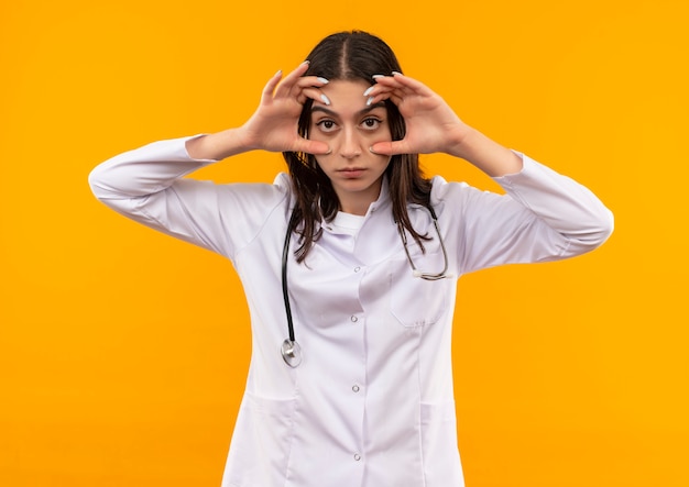 Joven doctora en bata blanca con estetoscopio alrededor de su cuello abriendo los ojos con los dedos sintiendo fatiga matutina de pie sobre la pared naranja