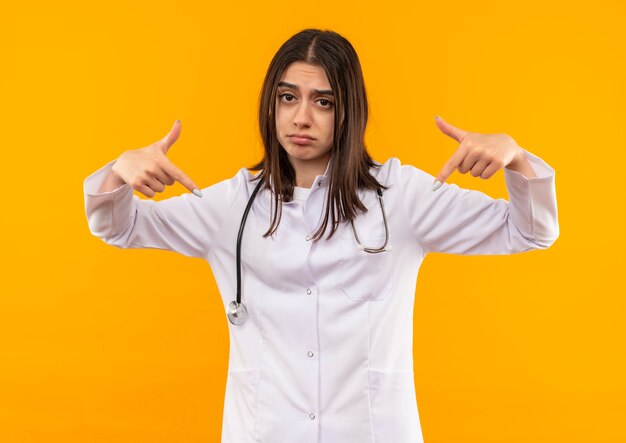 Joven doctora en bata blanca con estetoscopio alrededor de su cuello apuntando con el dedo índice a sí misma mirando confundida con expresión triste de pie sobre la pared naranja