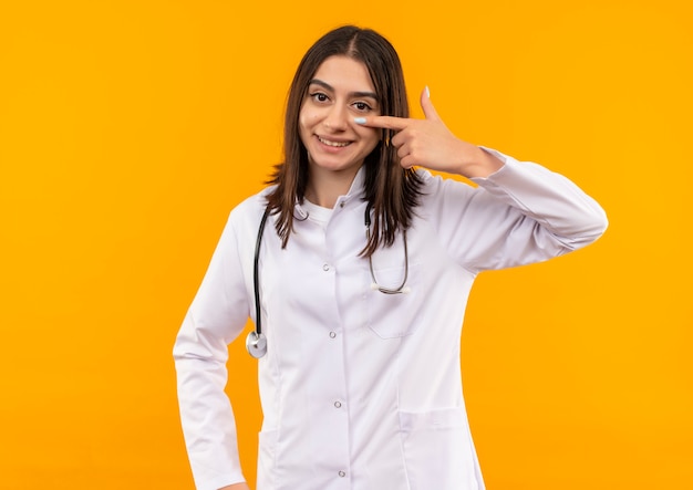 Joven doctora en bata blanca con estetoscopio alrededor de su cuello apuntando con el dedo a su ojo sonriendo de pie sobre la pared naranja