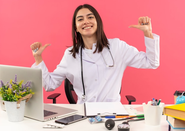 Foto gratuita joven doctora en bata blanca con estetoscopio alrededor de su cuello apuntando con los dedos a sí misma mirando satisfecha de sí misma sentada en la mesa con un portátil sobre una pared rosa