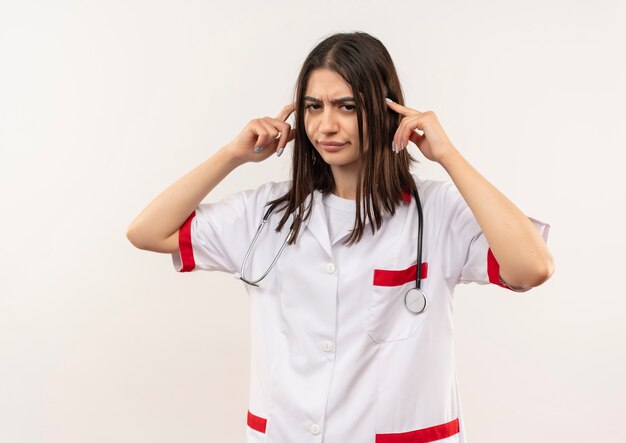 Joven doctora en bata blanca con estetoscopio alrededor de su cuello apuntando sus sienes con aspecto cansado y aburrido de pie sobre la pared blanca