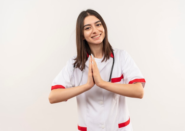 Joven doctora en bata blanca con estetoscopio alrededor de su cuello cogidos de la mano como gesto de namaste sintiéndose agradecido de pie sobre una pared blanca