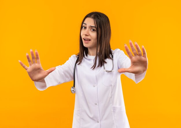 Joven doctora en bata blanca con estetoscopio alrededor de su cuello haciendo dejar de cantar sosteniendo las manos asustado de pie sobre la pared naranja