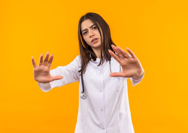 Joven doctora en bata blanca con estetoscopio alrededor de su cuello haciendo gesto de defensa con las manos mirando hacia el frente con cara seria de pie sobre la pared naranja