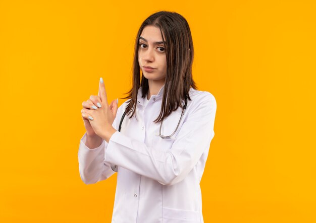 Joven doctora en bata blanca con estetoscopio alrededor de su cuello haciendo pistola o gesto de pistola con los dedos mirando hacia el frente con cara seria de pie sobre una pared naranja