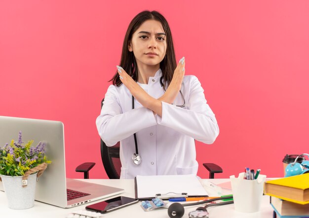 Joven doctora en bata blanca con estetoscopio alrededor de su cuello haciendo señal de stop con cara seria cruzando las manos sentado en la mesa con laptop sobre pared rosa