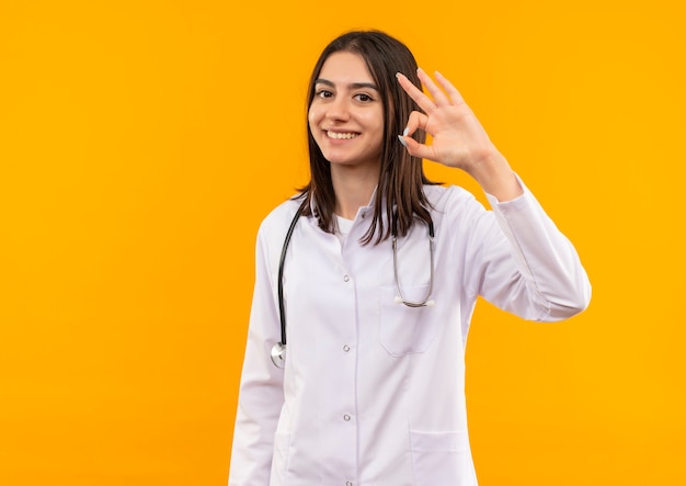 Joven doctora en bata blanca con estetoscopio alrededor de su cuello haciendo el signo de ok con los dedos sonriendo de pie sobre la pared naranja