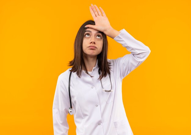 Joven doctora en bata blanca con estetoscopio alrededor de su cuello haciendo signo de perdedor sobre su cabeza mirando confundido parado sobre pared naranja