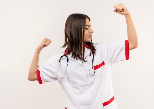 Joven doctora en bata blanca con estetoscopio alrededor de su cuello levantando la mano mostrando bíceps, concepto ganador de pie sobre una pared blanca