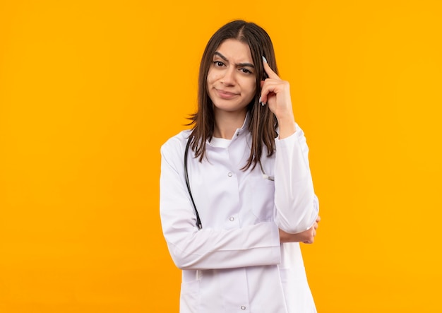 Joven doctora en bata blanca con estetoscopio alrededor de su cuello mirando al frente con expresión escéptica de pie sobre la pared naranja