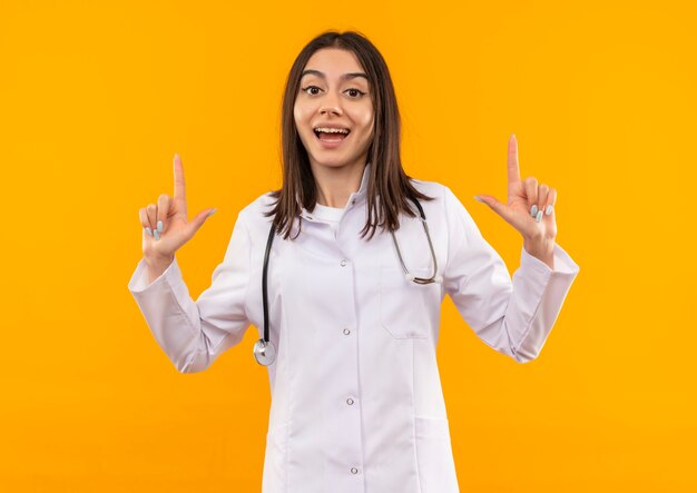 Joven doctora en bata blanca con estetoscopio alrededor de su cuello mirando al frente feliz y positivo sonriendo alegremente mostrando los dedos índices de pie sobre la pared naranja