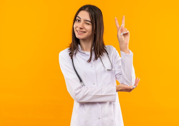 Joven doctora en bata blanca con estetoscopio alrededor de su cuello mirando al frente sonriendo y guiñando un ojo mostrando el signo de la victoria de pie sobre la pared naranja