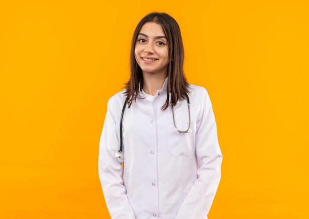 Joven doctora en bata blanca con estetoscopio alrededor de su cuello mirando al frente con una sonrisa de confianza de pie sobre la pared naranja