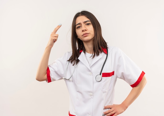 Joven doctora en bata blanca con estetoscopio alrededor de su cuello mirando hacia el frente disgustado mostrando dos dedos de pie sobre la pared blanca