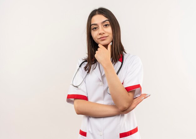 Joven doctora en bata blanca con estetoscopio alrededor de su cuello mirando hacia el frente con la mano en la barbilla mirando confiado pensando de pie sobre la pared blanca