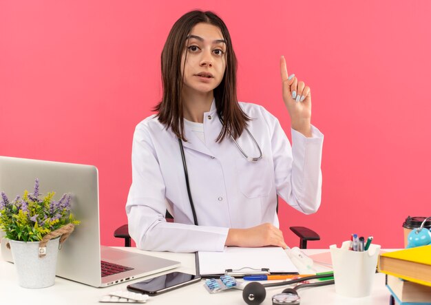 Joven doctora en bata blanca con estetoscopio alrededor de su cuello mirando hacia el frente mostrando el dedo índice con rostro serio sentado en la mesa con computadora portátil sobre pared rosa