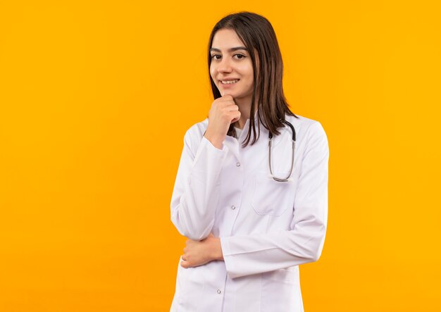 Joven doctora en bata blanca con estetoscopio alrededor de su cuello mirando hacia el frente perplejo parado sobre pared naranja