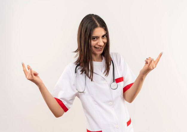 Joven doctora en bata blanca con estetoscopio alrededor de su cuello mirando hacia el frente sonriendo alegremente haciendo símbolo de rock con ambas manos de pie sobre la pared blanca