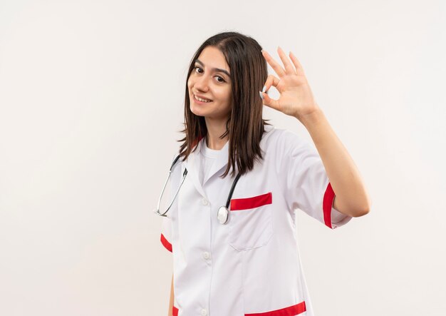 Joven doctora en bata blanca con estetoscopio alrededor de su cuello mirando hacia el frente sonriendo amigable mostrando signo ok de pie sobre una pared blanca