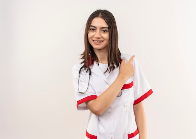 Joven doctora en bata blanca con estetoscopio alrededor de su cuello mirando hacia el frente sonriendo apuntando con el dedo hacia el lado de pie sobre la pared blanca