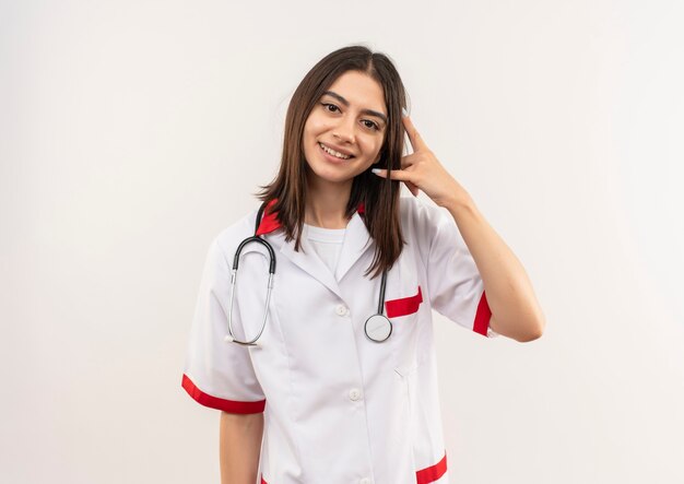 Joven doctora en bata blanca con estetoscopio alrededor de su cuello mirando hacia el frente sonriendo haciendo gesto de llamarme de pie sobre una pared blanca