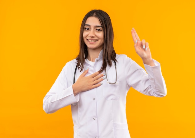 Joven doctora en bata blanca con estetoscopio alrededor de su cuello mirando hacia el frente tomando un juramento de pie sobre la pared naranja