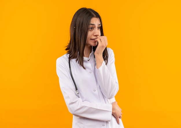 Joven doctora en bata blanca con estetoscopio alrededor de su cuello mirando a un lado estresado y nervioso parado sobre pared naranja