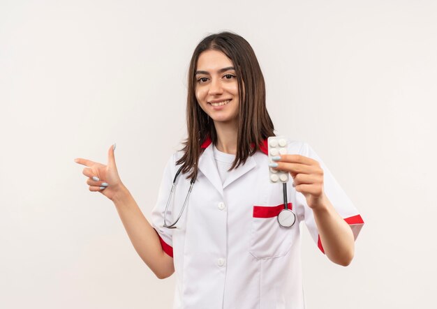 Joven doctora en bata blanca con estetoscopio alrededor de su cuello mostrando ampolla con pastillas apuntando con el dedo hacia el lado sonriendo de pie sobre la pared blanca