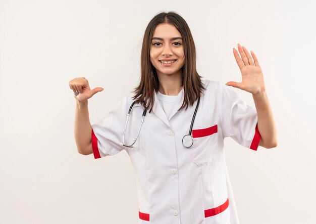 Joven doctora en bata blanca con estetoscopio alrededor de su cuello mostrando y apuntando hacia arriba con los dedos número seis sonriendo de pie sobre la pared blanca
