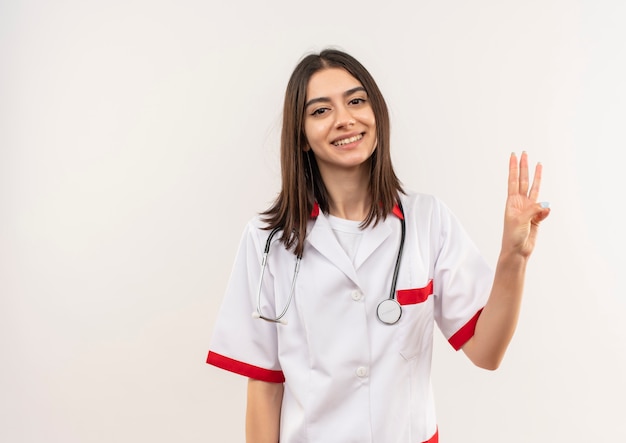 Joven doctora en bata blanca con estetoscopio alrededor de su cuello mostrando y apuntando hacia arriba con los dedos número tres sonriendo de pie sobre la pared blanca