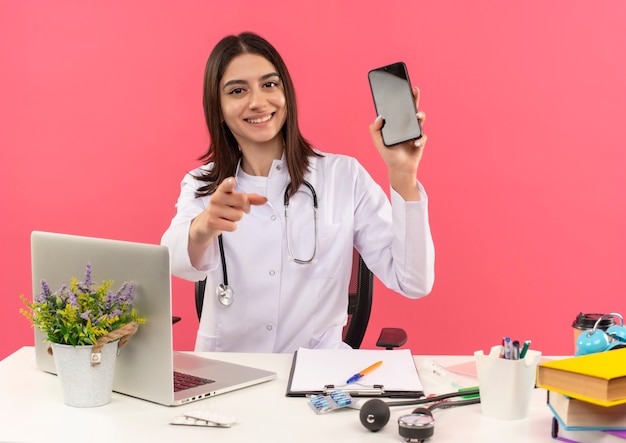 Joven doctora en bata blanca con estetoscopio alrededor de su cuello mostrando smartphone apuntando con el dedo al frente sentado en la mesa con laptop sobre pared rosa