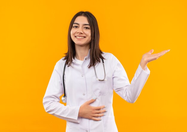 Joven doctora en bata blanca con estetoscopio alrededor de su cuello presentando algo con el brazo de su mano sonriendo alegremente de pie sobre la pared naranja