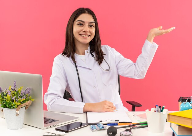 Joven doctora en bata blanca con estetoscopio alrededor de su cuello presentando algo con el brazo de su mano sonriendo sentado en la mesa con laptop sobre pared rosa