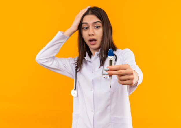 Joven doctora en bata blanca con estetoscopio alrededor de su cuello que muestra un termómetro digital mirándolo sorprendido de pie sobre la pared naranja