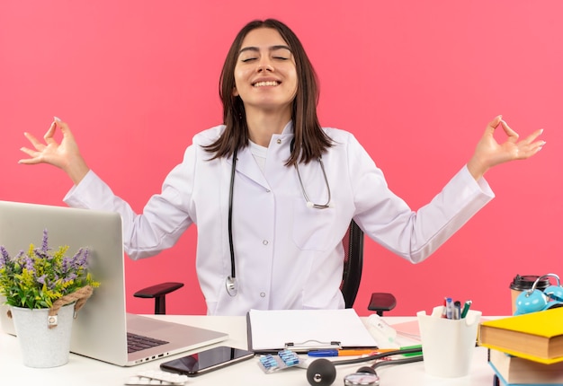 Joven doctora en bata blanca con estetoscopio alrededor de su cuello relajándose con los ojos cerrados haciendo un gesto de meditación con los dedos sentado en la mesa con una computadora portátil sobre una pared rosa