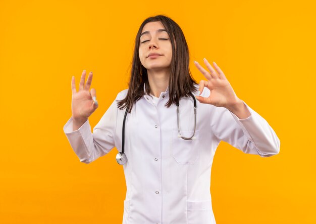 Joven doctora en bata blanca con estetoscopio alrededor de su cuello relajándose con los ojos cerrados haciendo gestos de meditación con los dedos de pie sobre la pared naranja