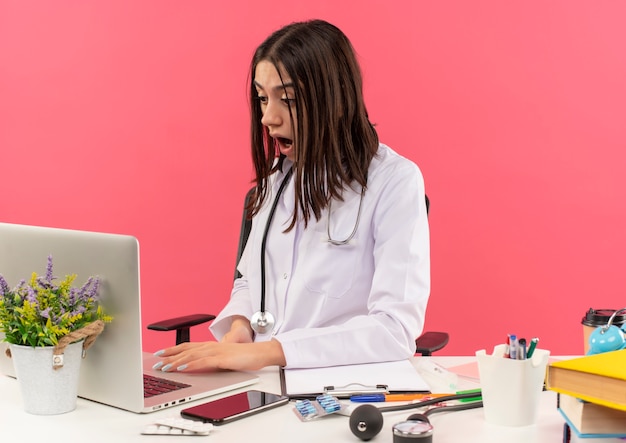 Joven doctora en bata blanca con estetoscopio alrededor de su cuello sentada en la mesa con laptop mirando confundida y sorprendida sobre pared rosa