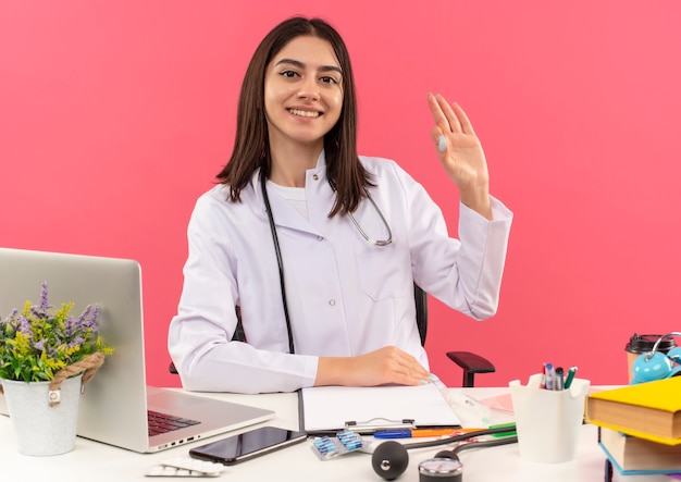 Joven doctora en bata blanca con estetoscopio alrededor de su cuello sonriendo mirando al frente mostrando signo de ok sentado en la mesa con laptop sobre pared rosa