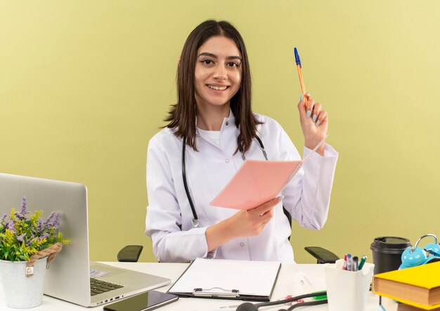 Joven doctora en bata blanca con estetoscopio alrededor de su cuello sosteniendo un cuaderno y un bolígrafo mirando al frente con una sonrisa en la cara sentada en la mesa con una computadora portátil sobre una pared clara