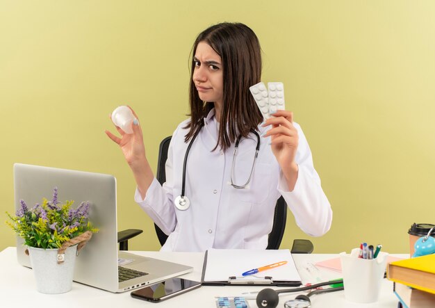 Joven doctora en bata blanca con estetoscopio alrededor de su cuello sosteniendo un frasco de prueba y una ampolla de píldoras con aspecto disgustado sentado en la mesa con una computadora portátil sobre una pared de luz