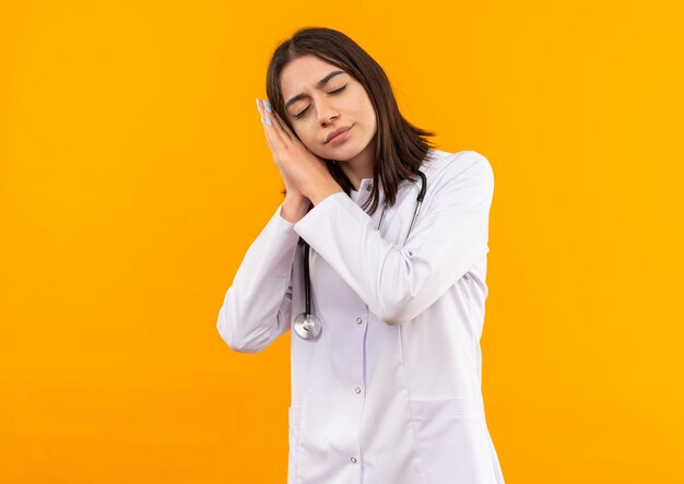 Joven doctora en bata blanca con estetoscopio alrededor de su cuello sosteniendo las palmas juntas inclinando la cabeza sobre las palmas quiere dormir con aspecto cansado de pie sobre la pared naranja