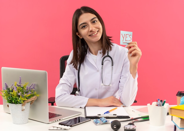 Joven doctora en bata blanca con estetoscopio alrededor de su cuello sosteniendo un papel recordatorio con la palabra sí mirando al frente con una sonrisa en la cara sentada en la mesa con una computadora portátil sobre una pared rosa