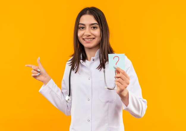 Joven doctora en bata blanca con estetoscopio alrededor de su cuello sosteniendo papel recordatorio con signo de interrogación apuntando con el dedo hacia el lado sonriendo mirando al frente parado sobre pared naranja