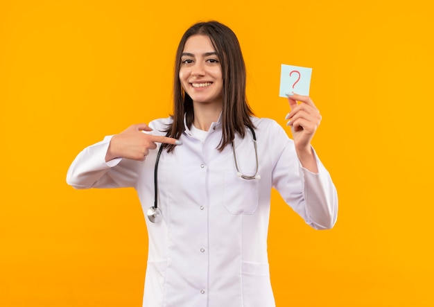 Joven doctora en bata blanca con estetoscopio alrededor de su cuello sosteniendo papel recordatorio con signo de interrogación apuntando con el dedo sonriendo mirando al frente parado sobre pared naranja