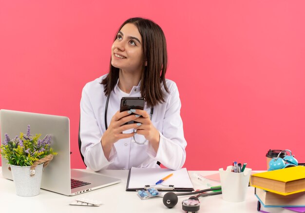 Joven doctora en bata blanca con estetoscopio alrededor de su cuello sosteniendo teléfono inteligente mirando a un lado sonriendo y pensando sentado en la mesa con computadora portátil sobre pared rosa
