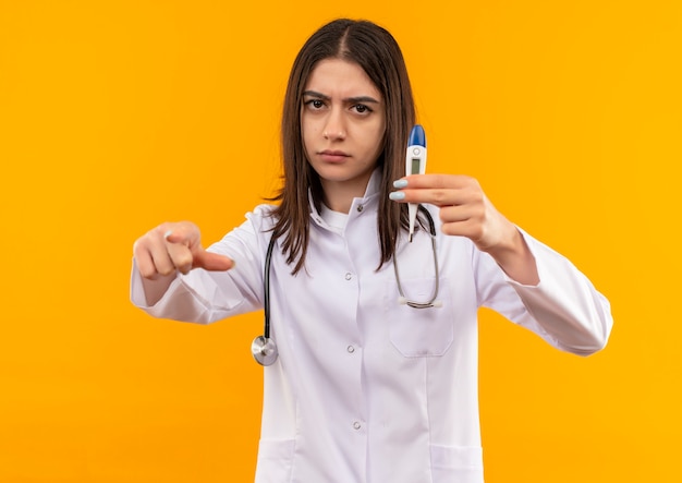 Joven doctora en bata blanca con estetoscopio alrededor de su cuello sosteniendo un termómetro digital apuntando con el dedo índice al frente con cara seria de pie sobre una pared naranja