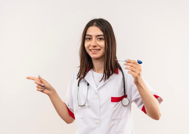 Joven doctora en bata blanca con estetoscopio alrededor de su cuello sosteniendo un termómetro digital apuntando con el dedo hacia el lado sonriendo de pie sobre la pared blanca