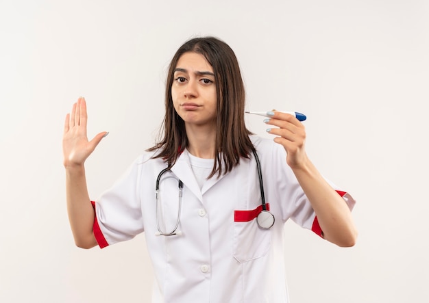 Joven doctora en bata blanca con estetoscopio alrededor de su cuello sosteniendo termómetro digital haciendo señal de stop con la mano con cara seria de pie sobre una pared blanca