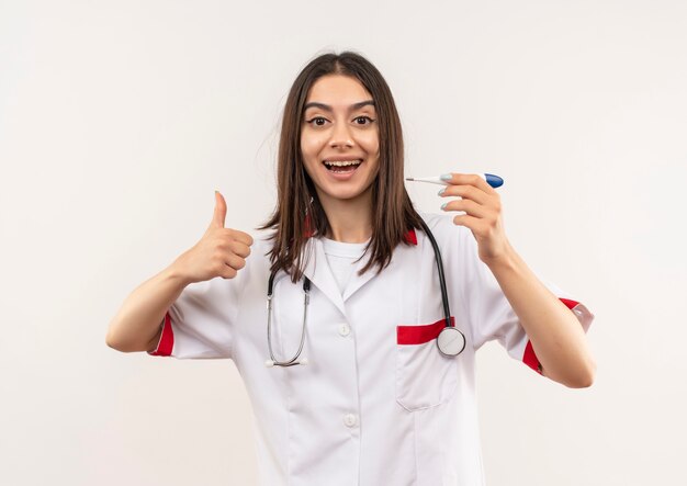 Joven doctora en bata blanca con estetoscopio alrededor de su cuello sosteniendo un termómetro digital sonriendo mostrando los pulgares para arriba sobre la pared blanca