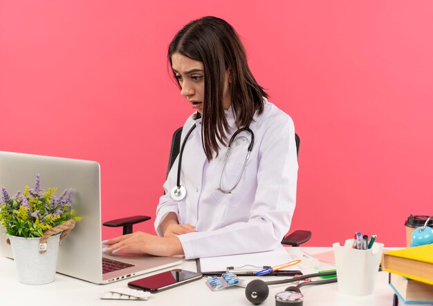 Joven doctora en bata blanca con estetoscopio alrededor de su cuello trabajando en una computadora portátil con aspecto confundido sentado en la mesa sobre la pared rosa